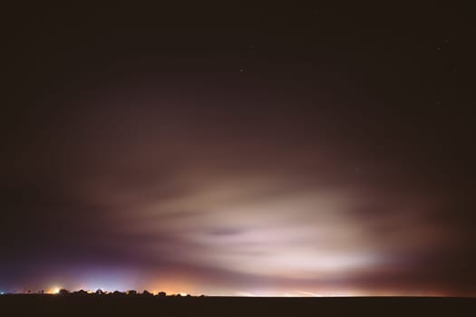Night Sky With Rare Glowing Stars Shining Through The Cloudiness Overcast Above Countryside Town Landscape. Rural Field Meadow In Early Spring.
