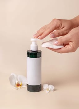 A woman hand pumping liquid on a make-up remover pad from a cosmetic bottle near orchid flowers on beige close up, label mockup. Skincare beauty product package, minimal composition..