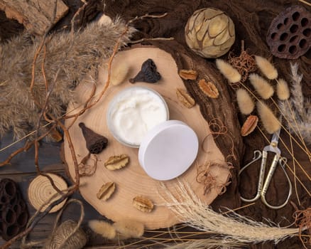 Opened cream jar with blank lid on wood near natural decorations top view, package mockup. Eco friendly skincare product, lotion or cream. Bohemian composition with dried leaves and flowers.