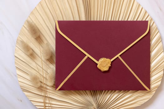Red envelope on dry beige palm leaf near hare's tail grass top view, mockup. Boho scene with envelope . Bohemian or close to nature greeting or wedding Invitation. Minimal composition.