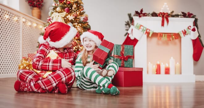 Brother and sister sitting on the floor indoors with Christmas gift boxes in their hands. Concept of the holiday eve and New Year.