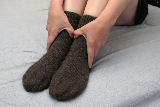 A young woman with slender legs sits on a bed at home in brown woolen socks. The concept of the world energy crisis.