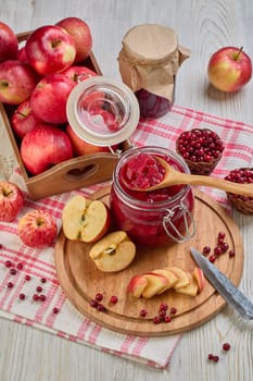 Still life with fresh lingonberries, red apples with knife on wooden cutting board and glass jar of homemade jam on light background. Autumn harvest, Home canning. Top view