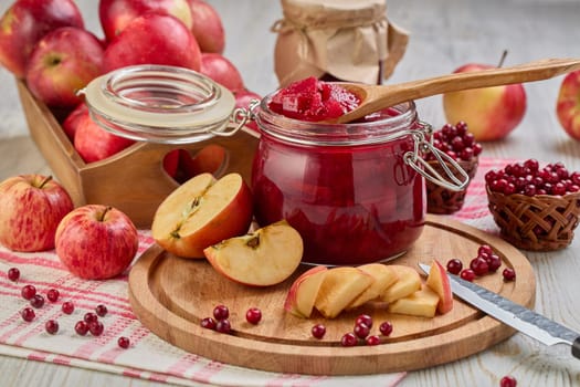 Still life with fresh lingonberries, red apples with knife on wooden cutting board and glass jar of homemade jam on light background. Autumn harvest, Home canning.