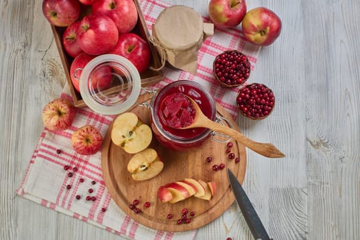 Wooden spoon with appetizing lingonberry and apple jam, jar of jam, fresh fruits and berries on light wooden table. Autumn harvest,  Home canning. Top view. Closeup