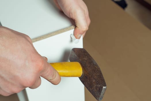 Male hands hold metal hammer with yellow plastic handle, driving dowel, assembling white drawer of dresser or wardrobe. Self-assembly of furniture at home, DIY, furniture repair and rearrangement