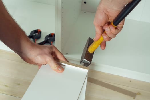 Male hands hold metal hammer with yellow plastic handle, driving dowel, assembling white drawer of dresser or wardrobe. Self-assembly of furniture at home, DIY, furniture repair and rearrangement