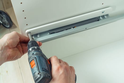 Male hands hold cordless electric screwdriver, screw to fix rail, assembling drawer of white dresser or wardrobe. Self-assembly of furniture at home, DIY, furniture repair and rearrangement