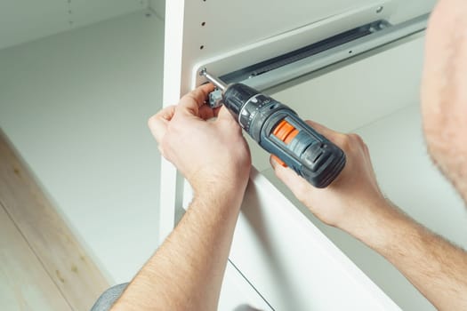 Male hands hold cordless electric screwdriver, screw to fix rail, assembling drawer of white dresser or wardrobe. Self-assembly of furniture at home, DIY, furniture repair and rearrangement