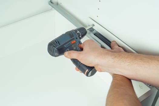 Male hands hold cordless electric screwdriver, screw to fix rail, assembling drawer of white dresser or wardrobe. Self-assembly of furniture at home, DIY, furniture repair and rearrangement