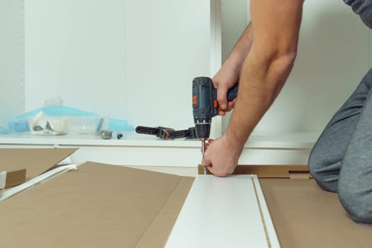 Male hands hold cordless electric screwdriver, screw to fix walls of white drawer, assembling dresser or wardrobe on floor. Self-assembly of furniture at home, DIY, furniture repair and rearrangement