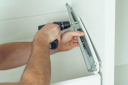 Male hands hold cordless electric screwdriver, screw to fix rail, assembling drawer of white dresser or wardrobe. Self-assembly of furniture at home, DIY, furniture repair and rearrangement