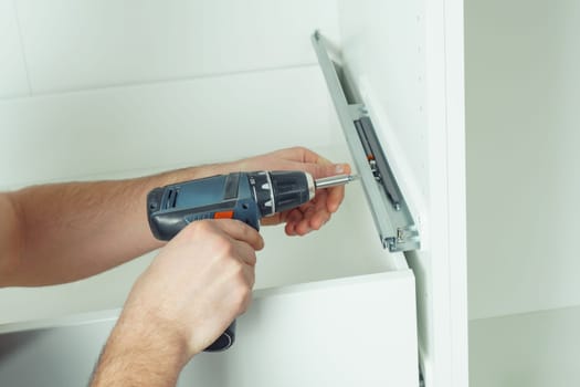 Male hands hold cordless electric screwdriver, screw to fix rail, assembling drawer of white dresser or wardrobe. Self-assembly of furniture at home, DIY, furniture repair and rearrangement
