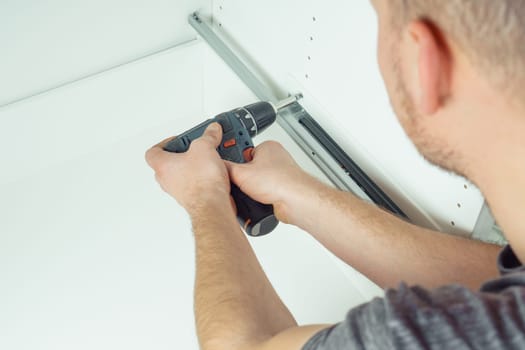 Male hands hold cordless electric screwdriver, screw to fix rail, assembling drawer of white dresser or wardrobe. Self-assembly of furniture at home, DIY, furniture repair and rearrangement
