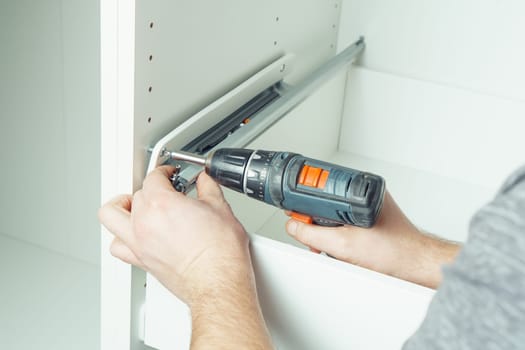 Male hands hold cordless electric screwdriver, screw to fix rail, assembling drawer of white dresser or wardrobe. Self-assembly of furniture at home, DIY, furniture repair and rearrangement