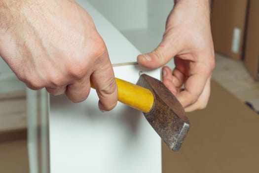 Male hands hold metal hammer with yellow plastic handle, driving dowel, assembling white drawer of dresser or wardrobe. Self-assembly of furniture at home, DIY, furniture repair and rearrangement