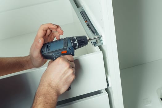 Male hands hold cordless electric screwdriver, screw to fix rail, assembling drawer of white dresser or wardrobe. Self-assembly of furniture at home, DIY, furniture repair and rearrangement