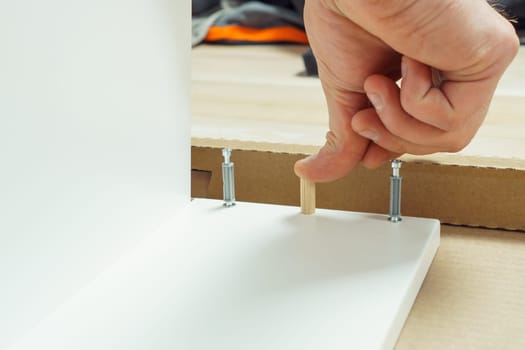 Male hands finger wooden dowel to fix walls of white drawer, assembling dresser or wardrobe on floor. Eccentric furniture ties on sides. Self-assembly of furniture at home, DIY, furniture repair