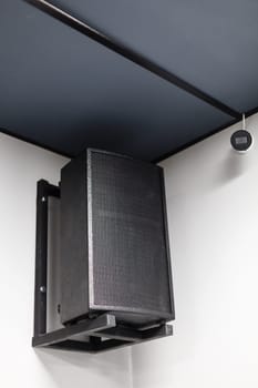 Close up of black big musical powerful speaker with protective grill hanging on wall in public place. Music speaker hangs on white wall near CCTV camera.