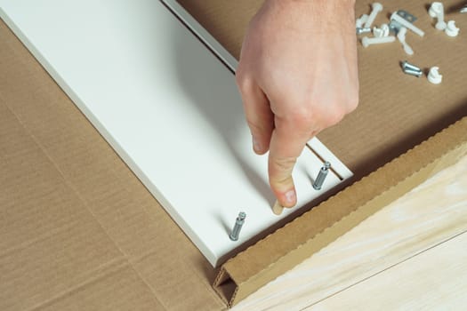 Male hands finger wooden dowel to fix walls of white drawer, assembling dresser or wardrobe on floor. Eccentric furniture ties on sides. Self-assembly of furniture at home, DIY, furniture repair