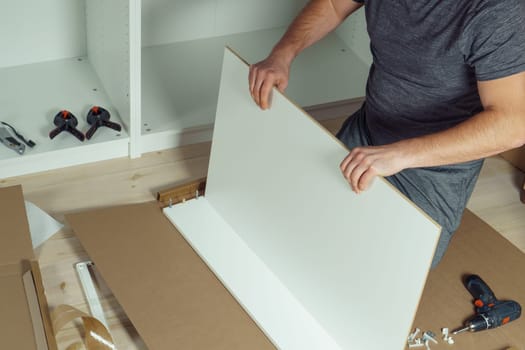 Male hands hold and insert bottom of white drawer, assembling dresser or wardrobe on floor. Electric screwdriver and clamps lie by side. Self-assembly of furniture at home, DIY, furniture repair