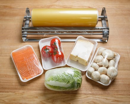 Tomatoes, mushrooms, cheese, salmon fillets and Chinese cabbage are wrapped in cling film for better storage. Roll of transparent food film for packing products on table near packed food portions.