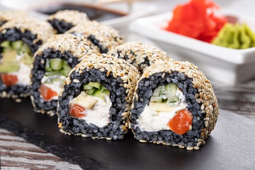 Close up of original unconventional sushi with black rice and covered with white sesame seeds. Close up of pieces of sushi roll with cheese, salmon and avocado on black metal board.