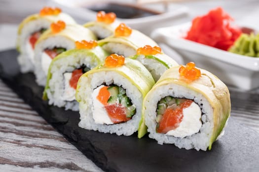Close up of delicious pieces of sushi covered with fresh slices of avocado and red caviar. Sushi roll with salmon, cheese and cucumber inside stands on black metal board for serving sushi.