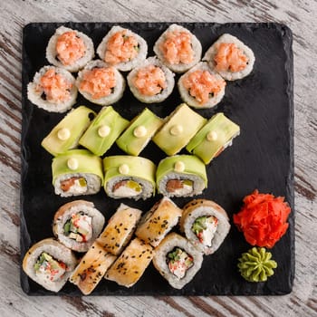 Asian cuisine. Large sushi set of various sushi rolls is served on black square metal slate. Top view of different rolls with variety of fresh ingredients and wasabi with ginger on edge.