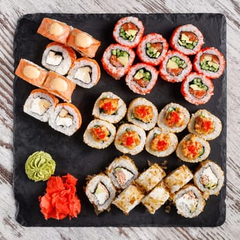 Asian cuisine. Large sushi set of various sushi rolls is served on black square metal slate. Top view of different rolls with variety of fresh ingredients and wasabi with ginger on edge.