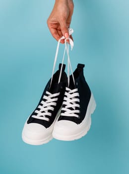 Female hand holding shoelaces stylish and fashionable model of black and white sneakers on blue background. Comfortable black sneakers with white soles and shoelaces for teens or children.