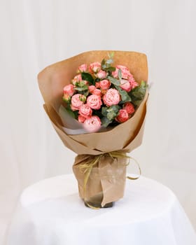 Bouquet of pink spray roses with sprigs of green eucalyptus in vase on stand on white textured background. Festive bouquet of flowers wrapped in brown kraft paper. Floral concept.