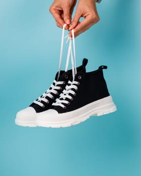 Female hand holding shoelaces stylish and fashionable model of black and white sneakers on blue background. Comfortable black sneakers with white soles and shoelaces for teens or children.