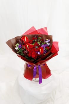 Beautiful large bouquet composed of red and purple flowers on stand on white fabric background. Bouquet of roses, calla lilies and eustoma wrapped in red floral film and tied with purple satin ribbon.