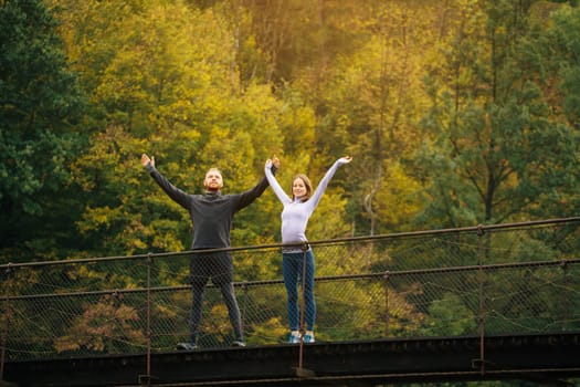 Athletic man and woman warm up before charging while walking in the mountains. Couple standing on a suspension bridge on a background of forest. Concept of tourist active life and adventure.