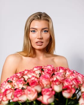 Spectacular blonde woman with makeup holds large number of pink roses and looks at camera on white background. Naked woman covering her body with large bouquet of small pink roses. Copy space.