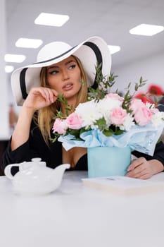 Fashion model portrait. Stylish beautiful young woman resting in restaurant sitting near bouquet of flowers. Woman with make-up dressed in white hat sits at table with teapot, cup and box with flowers.