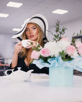 Portrait of beautiful elegant young woman sitting at table in cafe or restaurant. Young lady in white hat drinks tea sitting near bouquet of flowers. Selective focus.