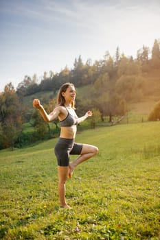 Young slender woman stands barefoot on one leg and practices balancing with yoga breathing exercises. Woman meditates in nature in a quiet mountain village. Concept of peace and meditation.