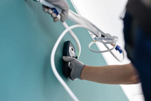 Male technician, electrician or builder uses modern laser length meter. Close up of man's hand in protective gloves attaching to wall between wires and cables digital measuring device.