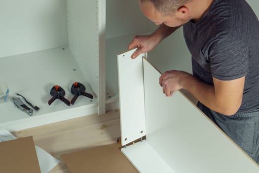 Male hands hold and insert bottom of white drawer, assembling dresser or wardrobe on floor. Electric screwdriver and clamps lie by side. Self-assembly of furniture at home, DIY, furniture repair