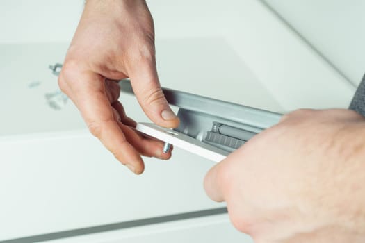 Male hands hold and insert metal screw to fix rail, assembling drawer of white dresser or wardrobe. Self-assembly of furniture at home, DIY, furniture repair and rearrangement
