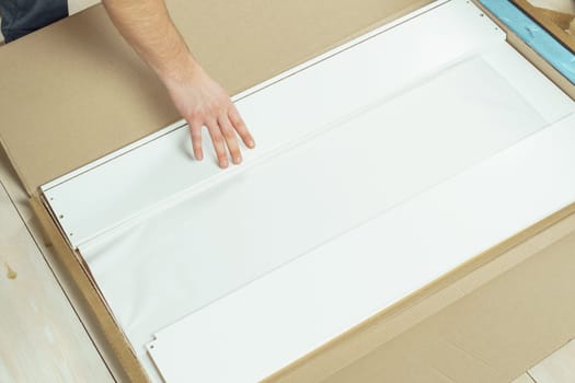 Male hands unpacking cardboard box with parts for assembling white chipboard drawer, dresser or wardrobe on floor. Self-assembly of furniture at home, DIY, furniture repair