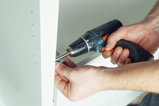Male hands hold cordless electric screwdriver, screw to fix rail, assembling drawer of white dresser or wardrobe. Self-assembly of furniture at home, DIY, furniture repair and rearrangement