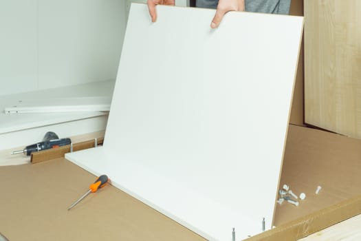 Male hands hold and insert bottom of white drawer, assembling dresser or wardrobe on floor. Electric screwdriver and screws lie by side. Self-assembly of furniture at home, DIY, furniture repair