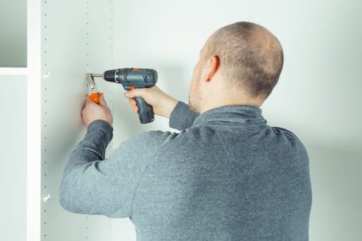 Male screw in to insert plastic support into one of series of holes to fix white shelf, assembling dresser or wardrobe. Holds screwdriver with pliers. Self-assembly of furniture, DIY, renovation
