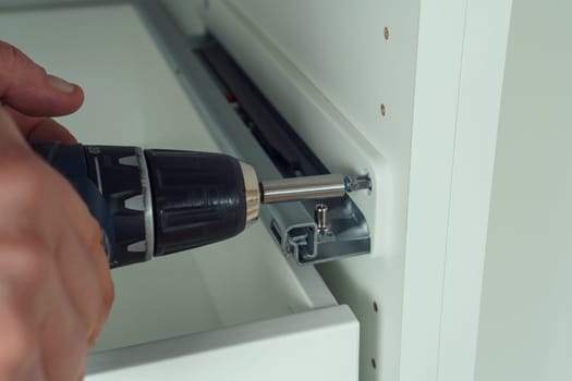 Male hands hold cordless electric screwdriver, screw to fix rail, assembling drawer of white dresser or wardrobe. Self-assembly of furniture at home, DIY, furniture repair and rearrangement