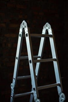 Construction. Metal folding step ladder in a dark room on a construction site. Close up of new construction step ladder on brick wall background.
