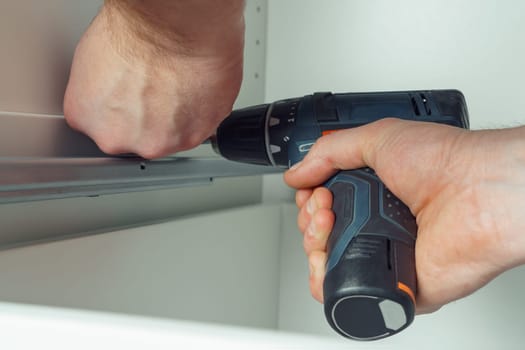 Male hands hold cordless electric screwdriver, screw to fix rail, assembling drawer of white dresser or wardrobe. Self-assembly of furniture at home, DIY, furniture repair and rearrangement