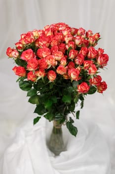 Bouquet consisting of many pink roses with tall stem and which stands on stand in glass vase. Festive bouquet of roses in transparent vase on background composed of white fabric.
